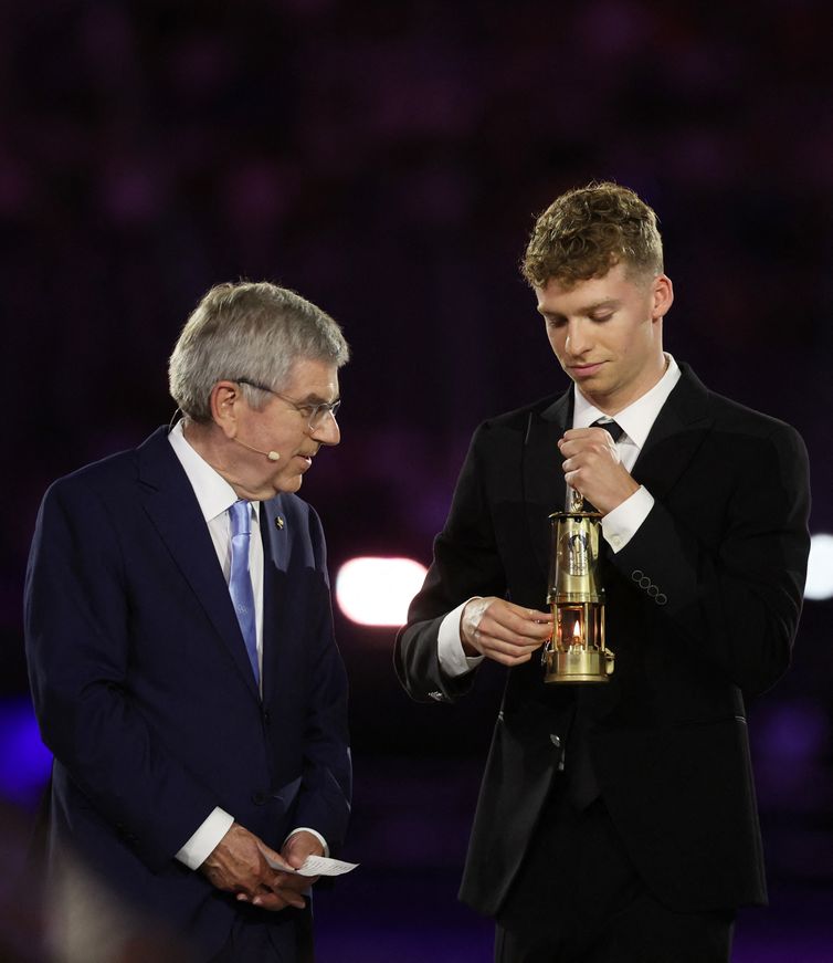 Reuters/Phil Noble/Direitos Reservados Paris 2024 Olympics - Ceremonies - Paris 2024 Closing Ceremony - Stade de France, Saint-Denis, France - August 11, 2024. Leon Marchand of France holds the Olympic flame as International Olympic Committee (IOC) President Thomas Bach looks on. REUTERS/Phil Noble